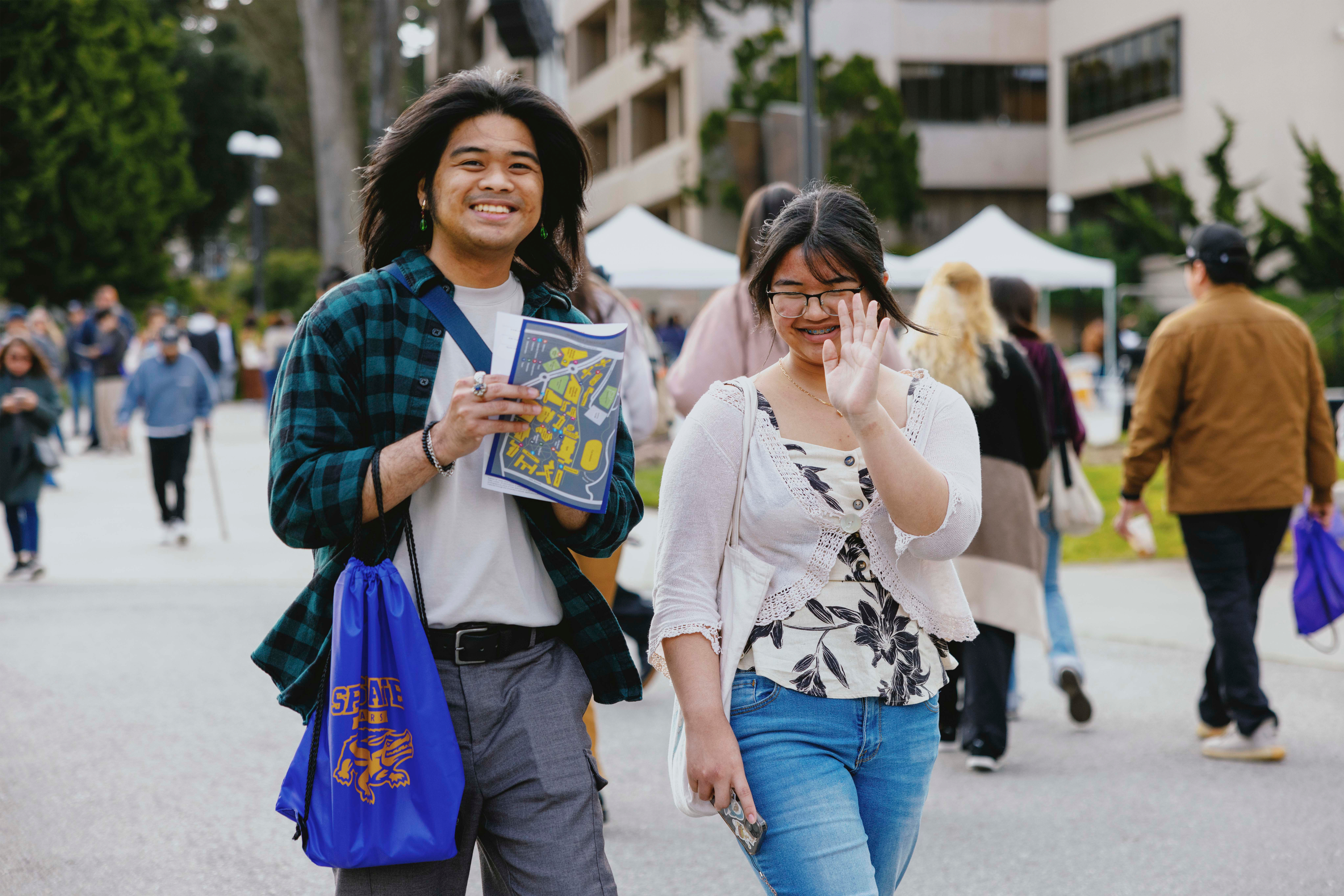 Two smiling prospective Students exploring sfsu. One student has a map in one hand and a sf state drawstring bag in the other. The other student puts their hand in front of their face to hide from the camera. A crowd of people and the administration building are in the background..