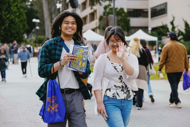 Two students walking on campus, smiling and waving at camera