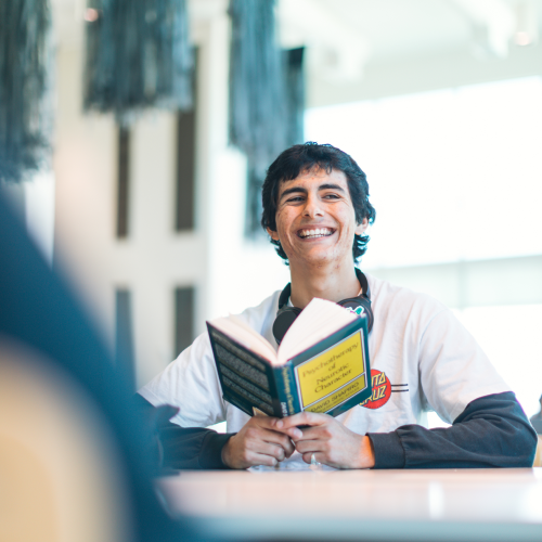student smiling while reading a book at a table